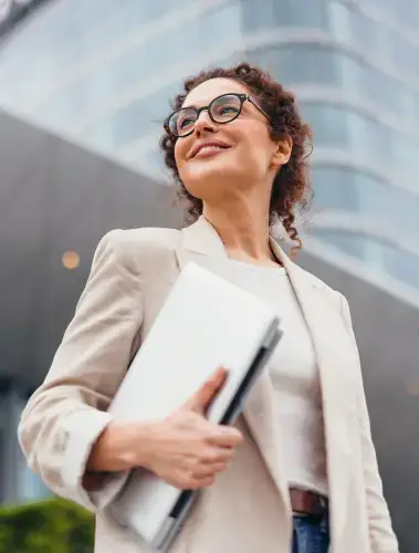 Woman in cream suit jacket and holding laptop under arm looking off to the distance and smiling for some reason